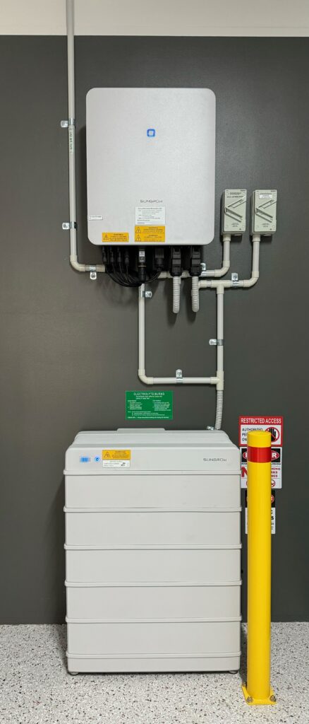 Solar battery and inverter setup. An inverter is at the top of the image. A multi-layer battery pack is stacked up from the floor, with a yellow bollard (including a red reflective warning strip) bolted into the floor to the right of the battery. The cables and conduits are neatly layered and everything is in front of dark-grey painted sheeting. The wall around the sheeting is painted white. The floor has a polyaspartic coating. There are a number of caution/warning labels stuck near the battery.