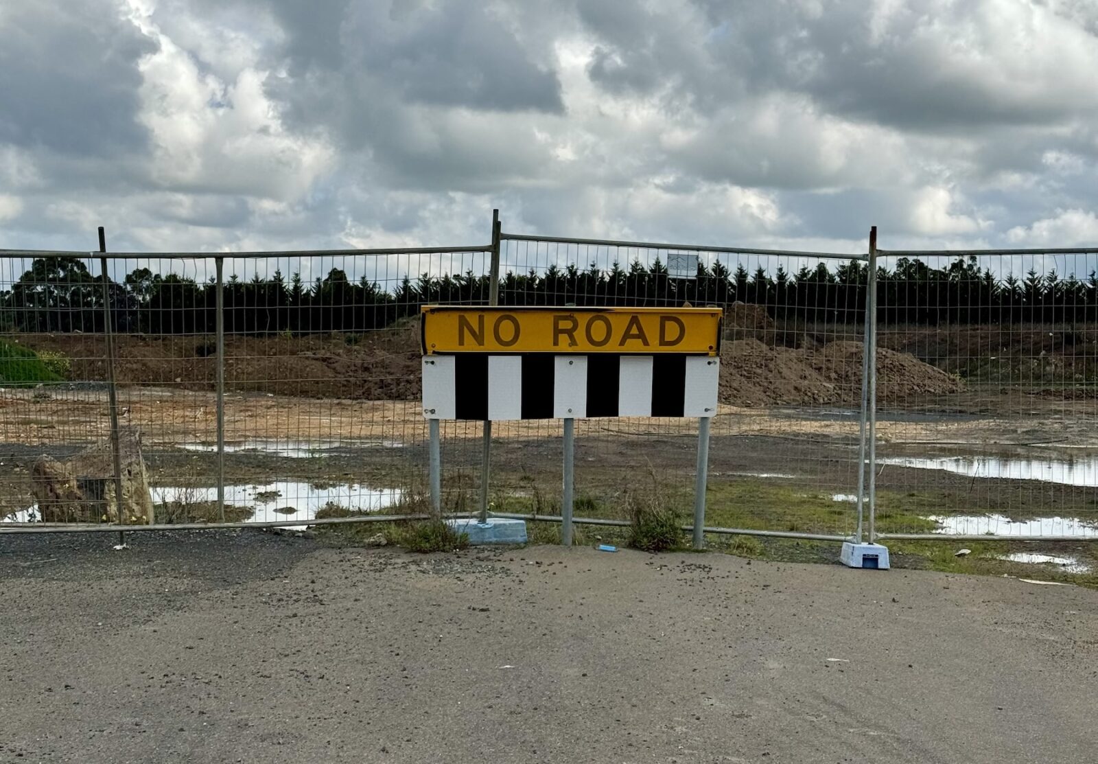 A road leading up to a fence, with a large sign reading "No Road". Beyond the fence is mnuddy, uncleared land.