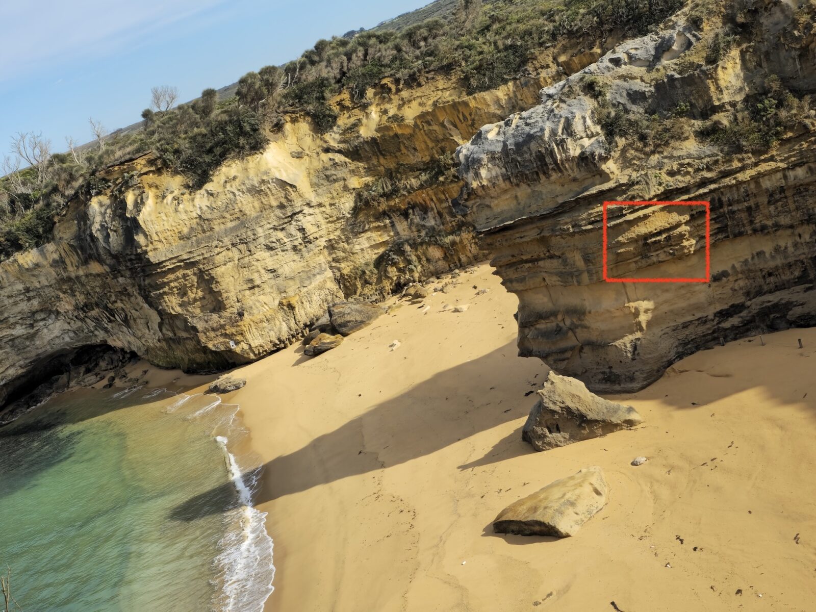 Beach at Loch Ard Gorge. Yellow sands, tall cliffs with shrubs on the top, and green/blue water. A section of a cliff has been highlighted with a red square.